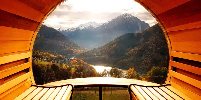 A scenic view from inside a wooden cabin or sauna, looking out through a large arched opening toward a mountain valley with a lake and autumn-colored trees.